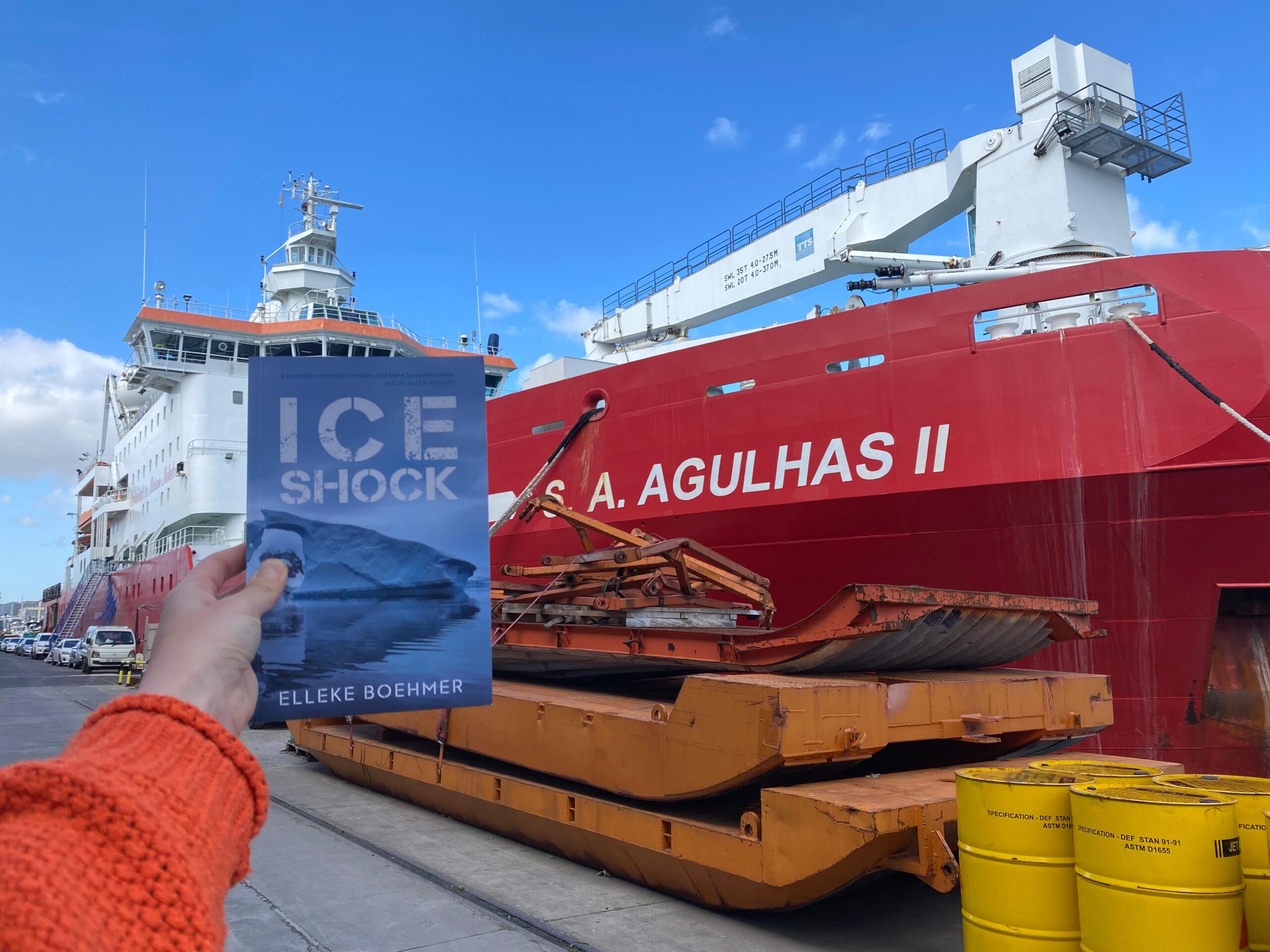 A hand holds up the novel Ice Shock in front of the ship S. A. Agulhas II